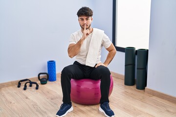 Hispanic man with beard sitting on pilate balls at yoga room asking to be quiet with finger on lips. silence and secret concept.