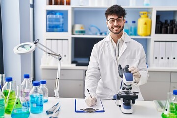 Young hispanic man scientist writing on document using microscope at laboratory