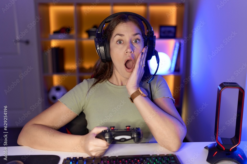 Canvas Prints Beautiful brunette woman playing video games wearing headphones afraid and shocked, surprise and amazed expression with hands on face