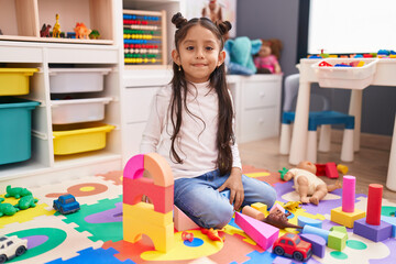 Adorable hispanic girl playing with construction blocks sitting on floor at kindergarten
