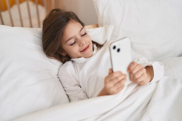 Adorable hispanic girl using smartphone lying on bed at bedroom