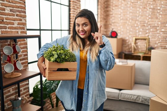 Young hispanic woman moving to a new home holding plants surprised with an idea or question pointing finger with happy face, number one