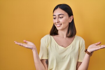 Hispanic girl wearing casual t shirt over yellow background smiling showing both hands open palms, presenting and advertising comparison and balance