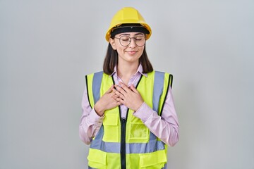 Hispanic girl wearing builder uniform and hardhat smiling with hands on chest with closed eyes and grateful gesture on face. health concept.