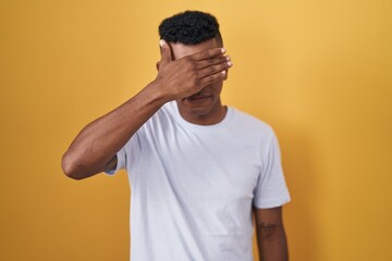Young hispanic man standing over yellow background covering eyes with hand, looking serious and sad. sightless, hiding and rejection concept