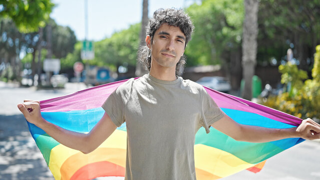 Young hispanic man holding rainbow flag with relaxed expression at street