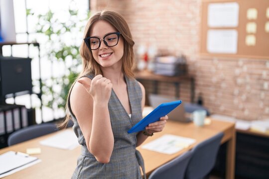 Caucasian woman working at the office wearing glasses smiling with happy face looking and pointing to the side with thumb up.