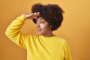 Fototapeta na wymiar Young african american woman standing over yellow background very happy and smiling looking far away with hand over head. searching concept.