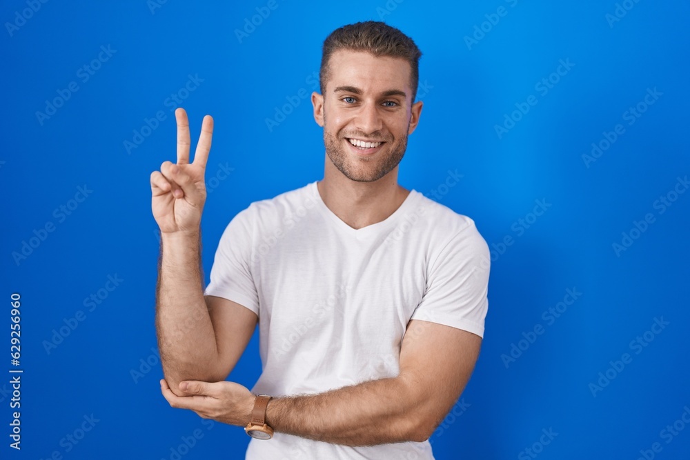 Sticker Young caucasian man standing over blue background smiling with happy face winking at the camera doing victory sign with fingers. number two.