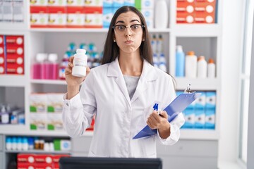 Young brunette woman working at pharmacy drugstore holding pills puffing cheeks with funny face. mouth inflated with air, catching air.