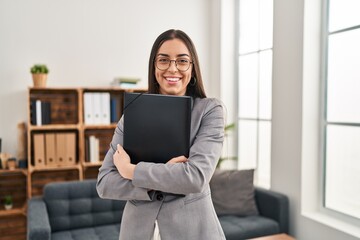 Young beautiful hispanic woman psychologist smiling confident holding binder at psychology clinic