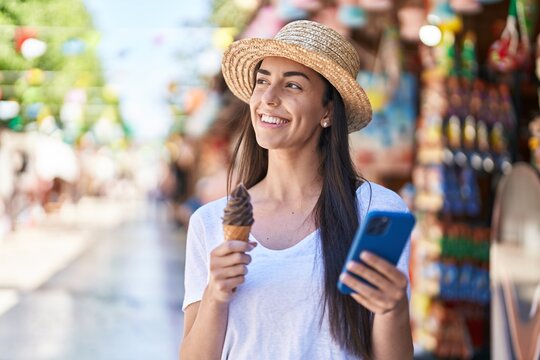 Young hispanic woman tourist using smartphone eating ice cream at street market