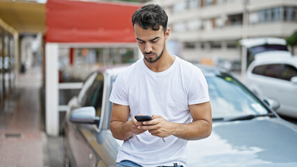 Young hispanic man using smartphone sitting on car at street