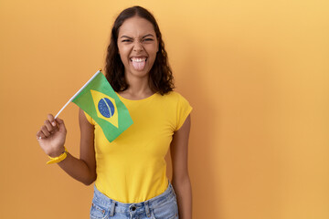 Young hispanic woman holding brazil flag sticking tongue out happy with funny expression. emotion...