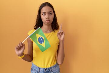 Young hispanic woman holding brazil flag pointing up looking sad and upset, indicating direction with fingers, unhappy and depressed.