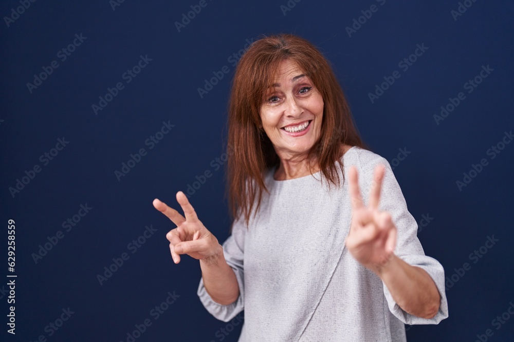 Sticker Middle age woman standing over blue background smiling looking to the camera showing fingers doing victory sign. number two.