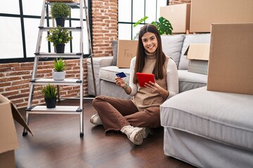 Young beautiful hispanic woman using touchpad and credit card sitting on floor at new home