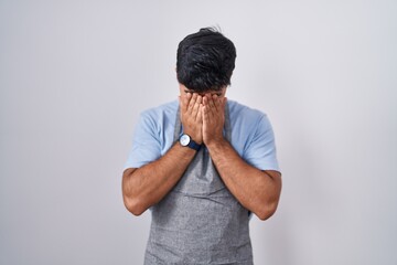 Hispanic young man wearing apron over white background with sad expression covering face with hands while crying. depression concept.