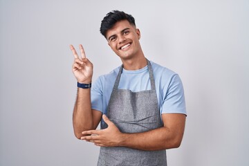 Hispanic young man wearing apron over white background smiling with happy face winking at the...