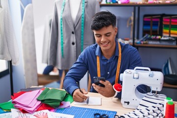Young hispanic man tailor using smartphone writing on notebook at clothing factory