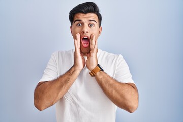 Hispanic man standing over blue background afraid and shocked, surprise and amazed expression with hands on face