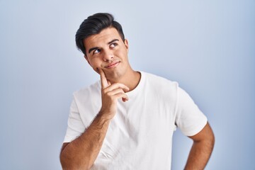 Hispanic man standing over blue background with hand on chin thinking about question, pensive expression. smiling with thoughtful face. doubt concept.
