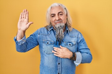 Middle age man with grey hair standing over yellow background swearing with hand on chest and open palm, making a loyalty promise oath