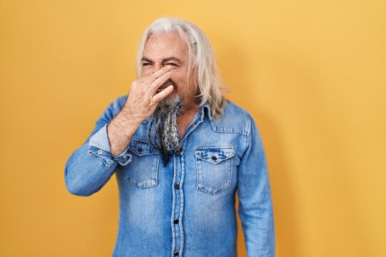 Middle Age Man With Grey Hair Standing Over Yellow Background Smelling Something Stinky And Disgusting, Intolerable Smell, Holding Breath With Fingers On Nose. Bad Smell