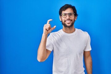 Fototapeta premium Handsome latin man standing over blue background smiling and confident gesturing with hand doing small size sign with fingers looking and the camera. measure concept.