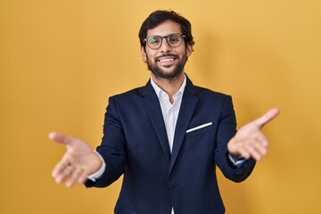 Handsome latin man standing over yellow background smiling cheerful offering hands giving assistance and acceptance.