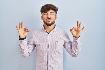 Arab man with beard standing over blue background relaxed and smiling with eyes closed doing meditation gesture with fingers. yoga concept.