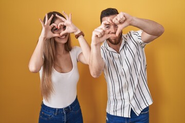 Young couple standing over yellow background doing heart shape with hand and fingers smiling looking through sign