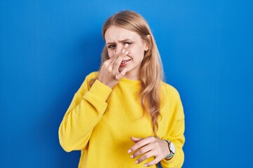 Young caucasian woman standing over blue background smelling something stinky and disgusting, intolerable smell, holding breath with fingers on nose. bad smell