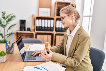 Young blonde woman business worker using laptop eating cereal bar at office
