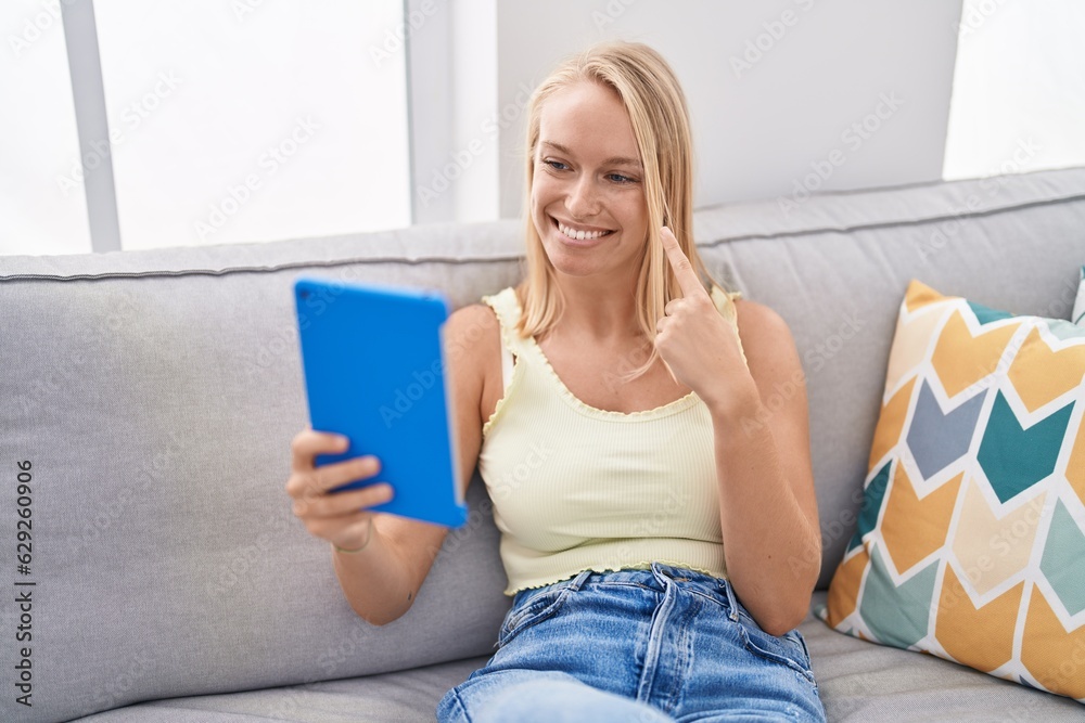 Sticker Young caucasian woman using touchpad sitting on the sofa smiling happy pointing with hand and finger