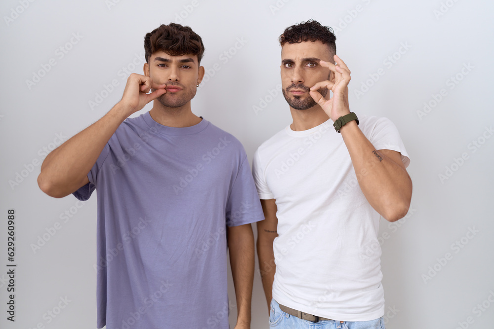 Poster homosexual gay couple standing over white background mouth and lips shut as zip with fingers. secret