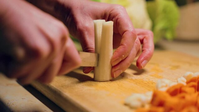 Woman with kitchen knife chopping raw white carrot on wooden cutting board at home