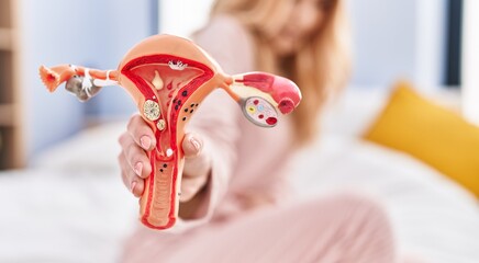 Young blonde woman suffering for menstrual pain holding anatomical model of uterus at bedroom