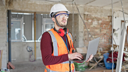 Young hispanic man builder using laptop looking around at construction site
