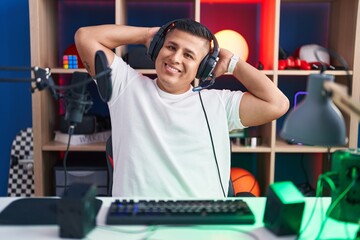 Young hispanic man playing video games relaxing and stretching, arms and hands behind head and neck...