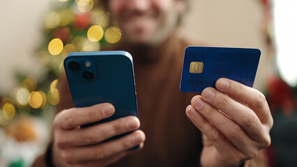 Young hispanic man using smartphone and credit card sitting on sofa by christmas tree at home
