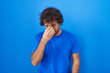 Hispanic young man standing over blue background tired rubbing nose and eyes feeling fatigue and headache. stress and frustration concept.