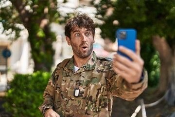 Hispanic young man wearing camouflage army uniform doing video call scared and amazed with open mouth for surprise, disbelief face