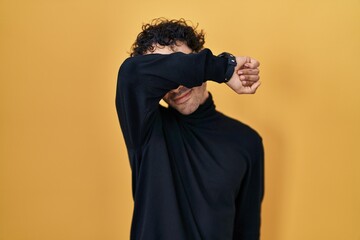 Hispanic man standing over yellow background covering eyes with arm smiling cheerful and funny. blind concept.