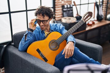 Young hispanic man musician playing classical guitar talking on smartphone at music studio
