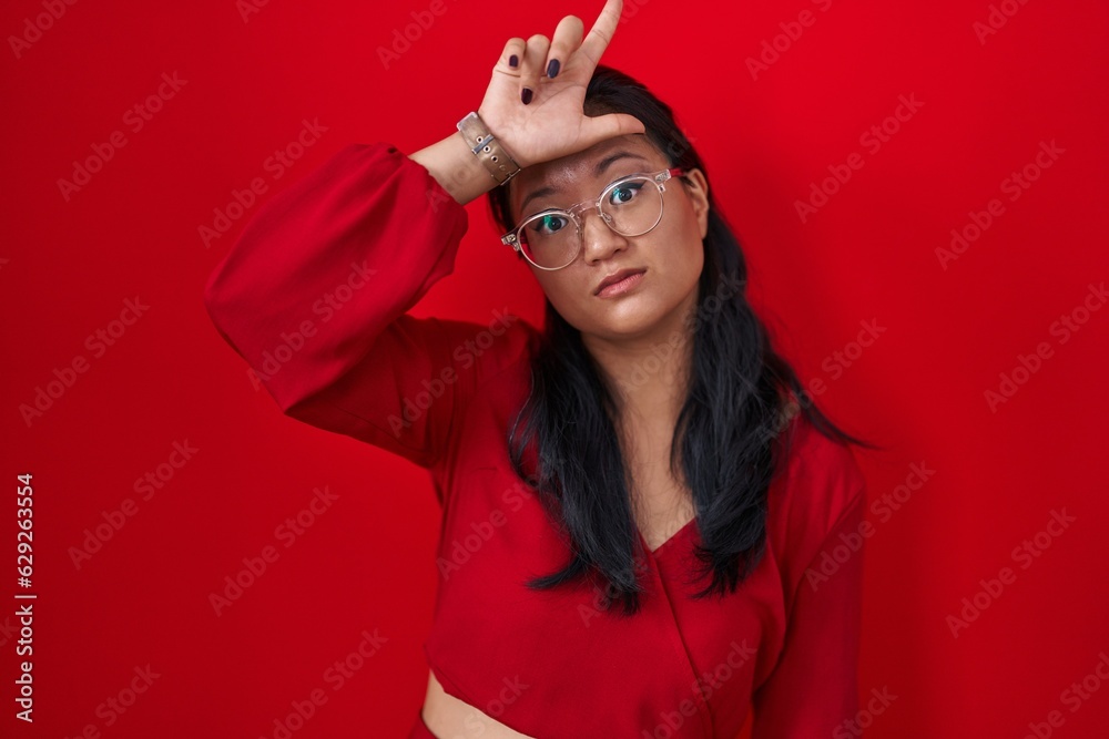 Poster asian young woman standing over red background making fun of people with fingers on forehead doing l