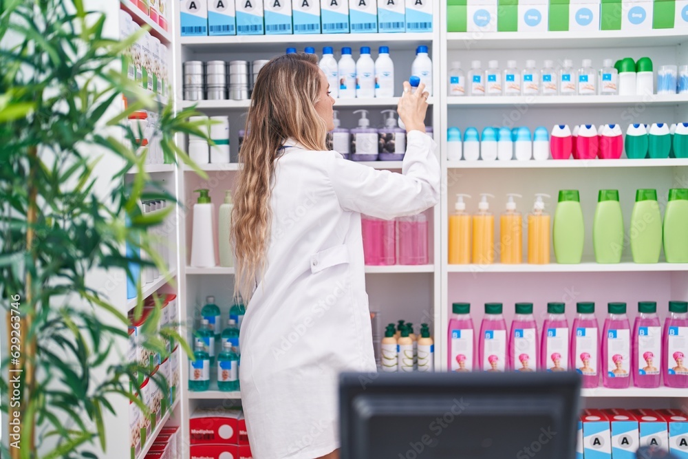 Wall mural Young blonde woman pharmacist holding product on shelving at pharmacy