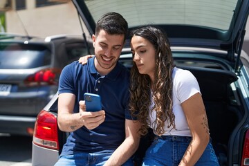 Fototapeta na wymiar Young hispanic couple sitting on car trunk using smartphone at street
