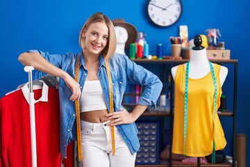 Young blonde woman tailor smiling confident leaning on clothes rack at sewing studio