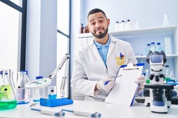 Hispanic man working at scientist laboratory holding blank clipboard smiling looking to the side and staring away thinking.